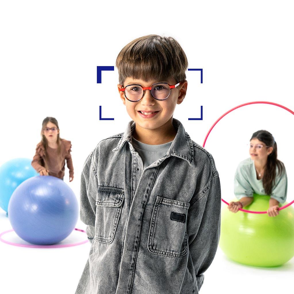 Brown-haired boy with round glasses wearing ZEISS MyoCare lenses stands in the foreground and smiles at the camera. In the background are two girls wearing ZEISS MyoCare lenses playing with hoops and gymnastic balls.