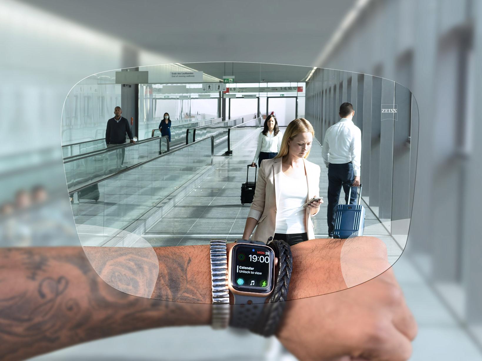 透過蔡司 SmartLife 數碼鏡片看手錶的人。在背景中可以看到典型的機場情景，人們推著行李箱上下扶梯。 