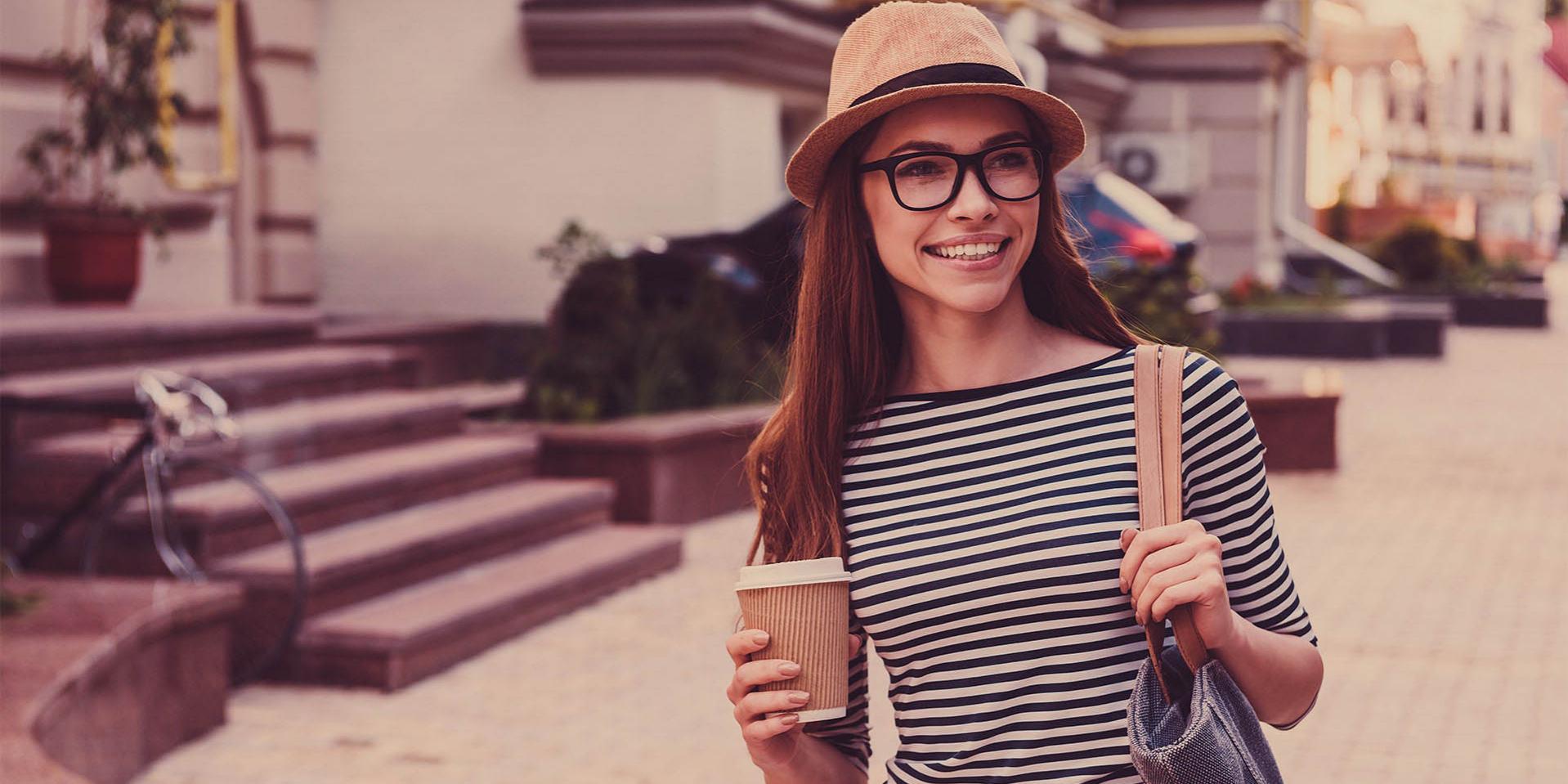 一位戴著帽子和眼鏡的年輕女子左肩挎著一個咖啡杯（右側）和一個包