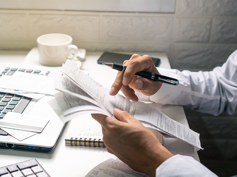 Typical office scene in which a person is busy with reviewing spreadsheets and documents. Only the arms of the person are visible, holding a stack of spreadsheets in his hands. In the background on the desk is a laptop. At the top left of the image is a pop up which shows a male person. The person has grey hair and beard, wear ZEISS Office lenses and has a neutral facial expression.