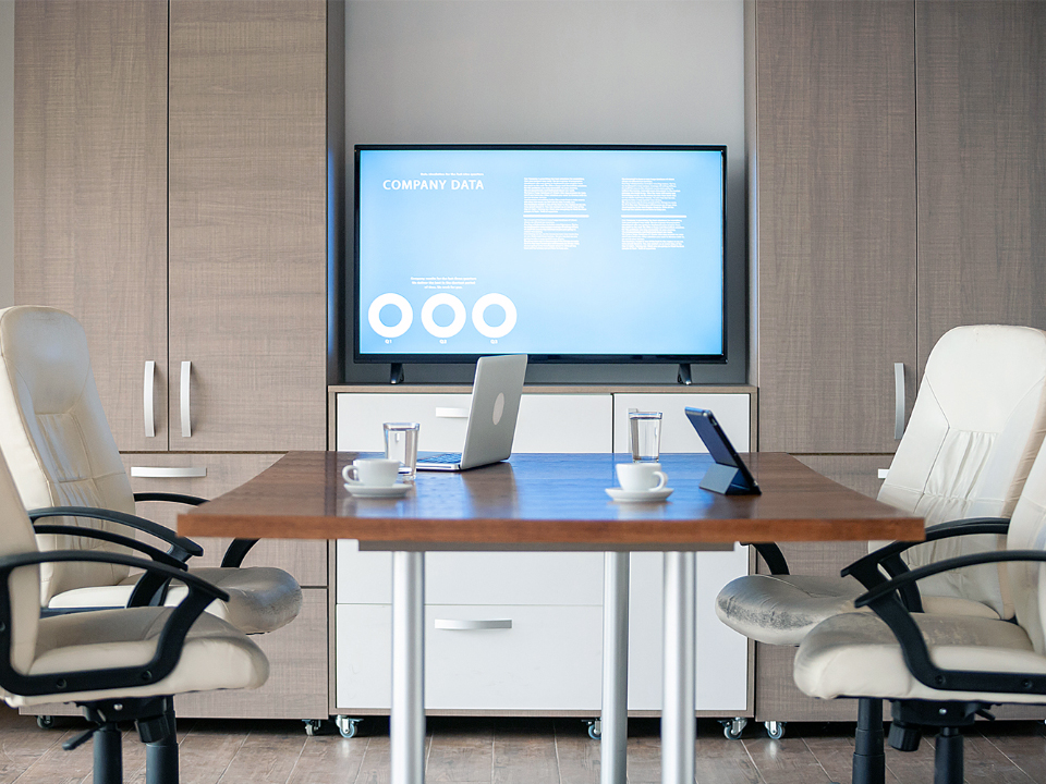 View from the desk of an executive office. A monitor is mounted on a wall a few meters away. There’s a small meeting table with office chairs and laptops on the table.