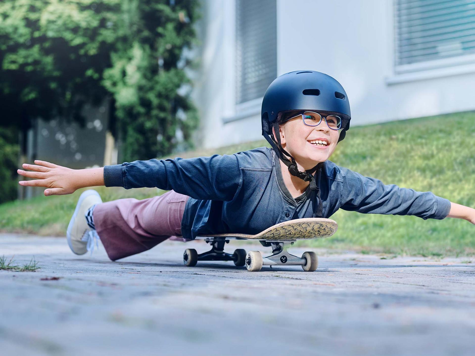 一個戴著頭盔和眼鏡的女孩躺在滑板上滑行，同時伸展雙臂。