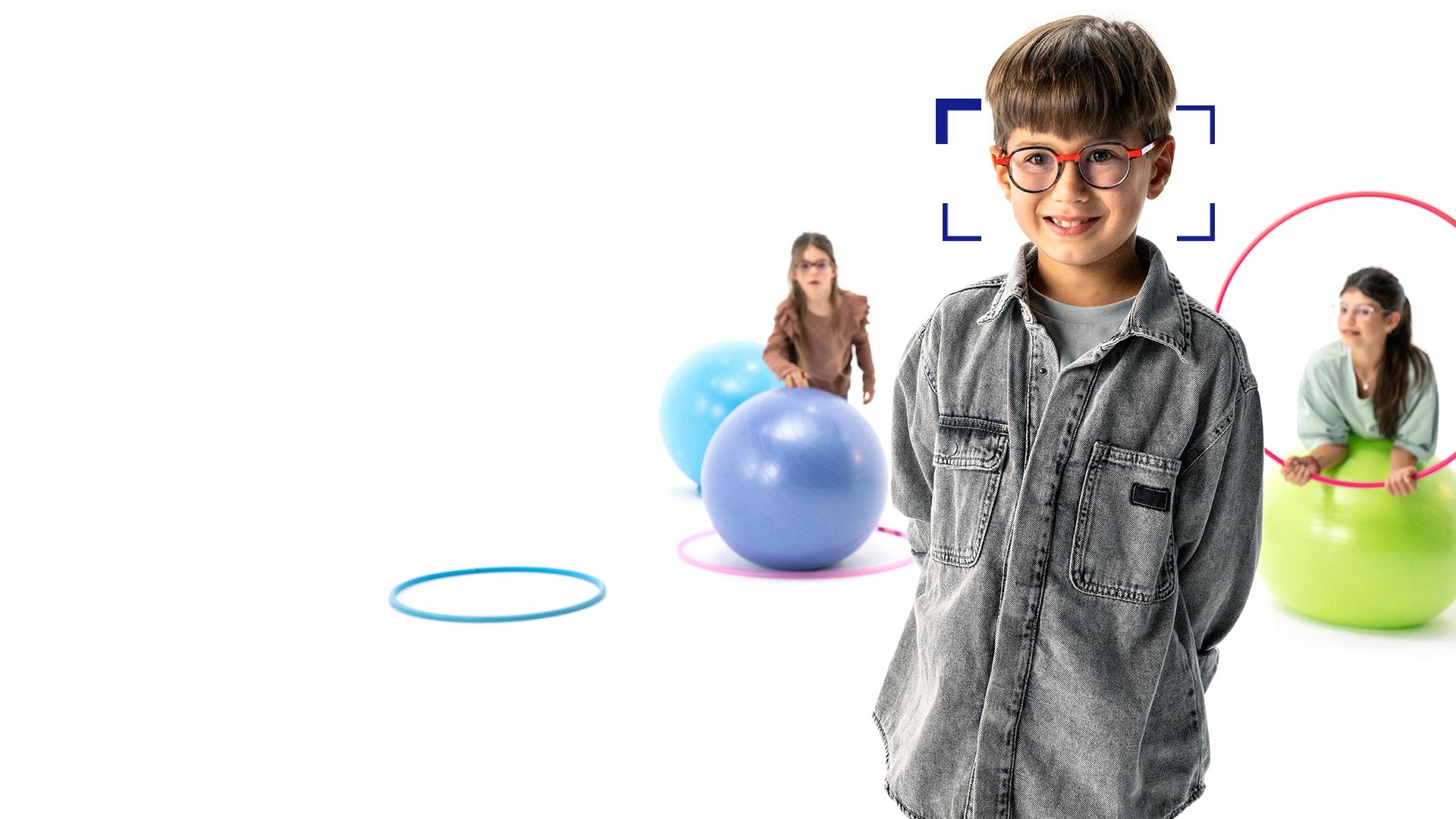 戴圓形眼鏡並使用蔡司 MyoCare 鏡片的棕髮男孩，站在前方對著相機燦笑。背景是兩位配戴蔡司MyoCare鏡片的女孩在玩體操圈和體操球。