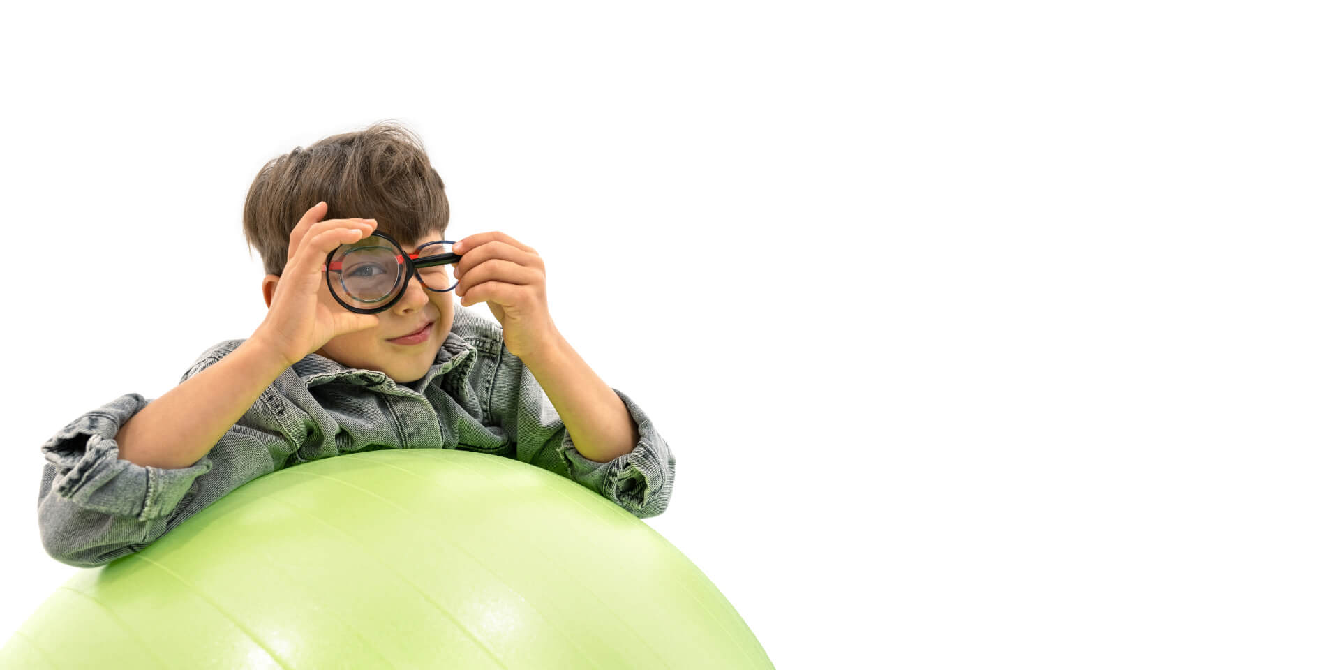 一個戴著蔡司近視控制眼鏡的男孩倚靠在體操球上，拿著放大鏡，單眼透過放大鏡觀看。