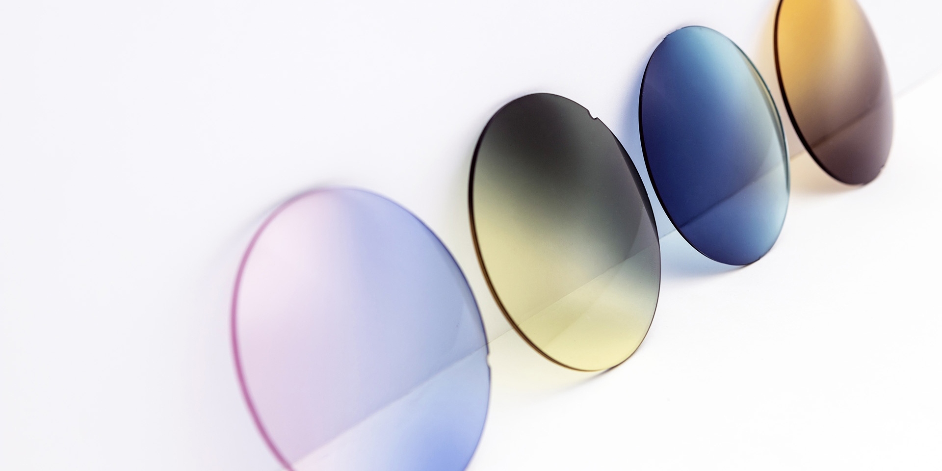放置於白色平面上的各種染色太陽鏡鏡片：粉紫色、黃灰色、漸變藍色和漸變啡色。
